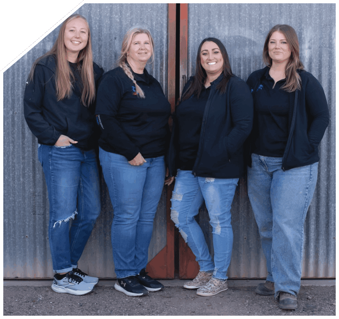 Four women standing casually against a wooden wall, dressed in black tops and jeans.