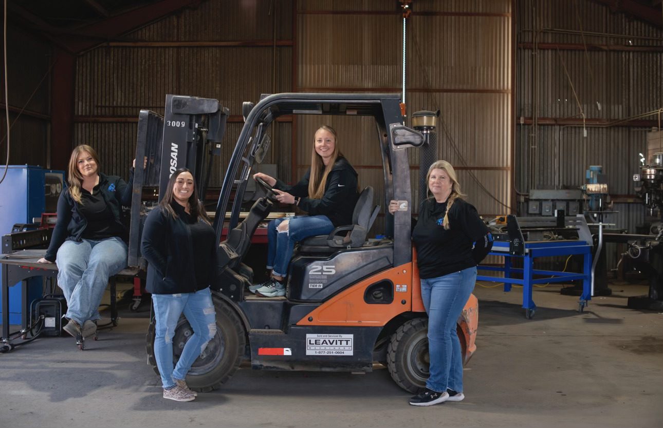 Three women posing with a forklift inside a warehouse.